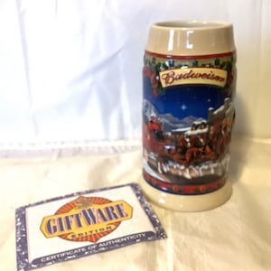 Può includere: Una boccale di birra natalizia Budweiser in ceramica con una scena invernale che presenta un carro rosso trainato da cavalli. Il boccale ha uno sfondo beige con un bordo rosso e verde. Il testo "Budweiser" è stampato su uno stendardo bianco sul boccale. Il boccale viene fornito con un certificato di autenticità.