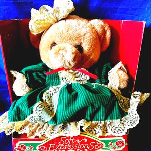 May include: A soft teddy bear with light brown fur, wearing a green dress with white lace trim, and a yellow bow. The bear is in a red box with the text "Soft Expressions".