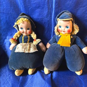 May include: Two vintage Dutch dolls with blue velvet outfits and bonnets. One doll has blonde braids and a floral apron. The other doll has a yellow scarf. Both dolls have rosy cheeks and yellow shoes.