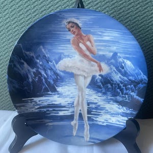May include: A porcelain plate featuring a ballerina in a white tutu, standing on a blue and white water scene with mountains in the background.