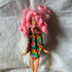 May include: A vintage doll with bright pink curly hair and blue eyes. The doll wears a colorful patterned jacket with red, green, and yellow accents. The doll is wearing red shoes.
