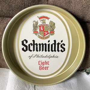 May include: A gold-colored metal tray with a white center featuring the logo for Schmidt's of Philadelphia Light Beer. The logo includes a crest with a lion and the text "Since 1860".