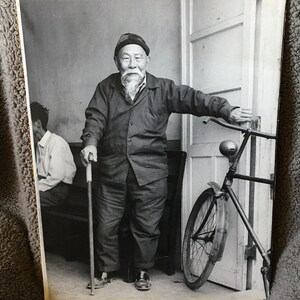 May include: A black and white photograph of an older man with a long white beard, wearing a dark jacket and a cap, standing with a cane next to a bicycle. The man is looking at the camera and smiling.