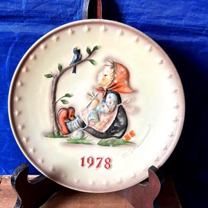 May include: A decorative ceramic plate featuring a seated child knitting, with a bird perched on a tree branch. The plate has a cream-colored background with a star-studded border and the year "1978" in red.