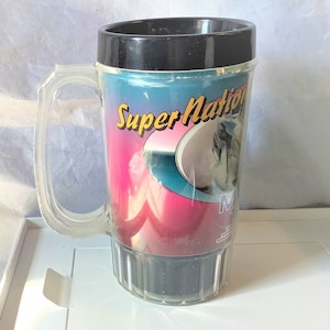 Può includere: Una tazza di plastica trasparente con un coperchio e un manico neri. La tazza ha un design colorato con le parole "Super Nation" e una grafica di una persona che fa surf.