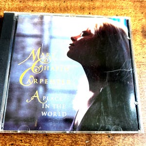 May include: A CD case featuring the album "A Place in the World" by Mary Chapin Carpenter. The cover art shows a profile of a woman's face in silhouette, with the artist's name and album title in gold lettering.