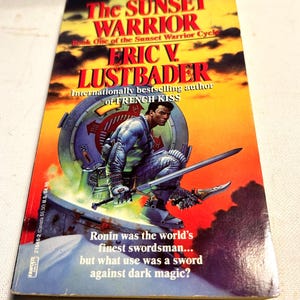 Puede incluir: Un libro de bolsillo titulado "The Sunset Warrior" de Eric V. Lustbader. La portada presenta a un guerrero con una espada sobre un fondo de puesta de sol y un diseño futurista. El texto dice: "Ronin era el mejor espadachín del mundo... pero ¿de qué servía una espada contra la magia oscura?"