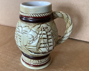May include: A ceramic beer stein with a rope-like handle. The stein features a raised design of a ship sailing into a harbor with buildings in the background. The text "Genuine Bark & Stay Sail" is printed on the bottom of the stein.