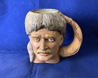 May include: A ceramic mug shaped like a man's head wearing a fur hat. The mug has a brown handle shaped like a horn.