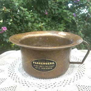 May include: A round, weathered brass chamber pot with a single side handle. An oval black label on the front features white text reading: NOTICE TO PASSENGERS. DO NOT EMPTY THIS TOILET OUT OF TRAIN WINDOW. CENTRAL PACIFIC R.R.