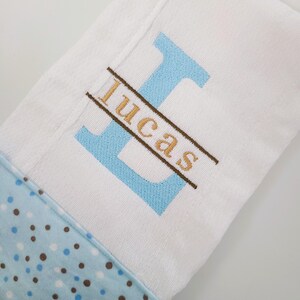 May include: White baby blanket with a blue polka dot border. The blanket features a large, light blue embroidered letter "L" with the name "Lucas" embroidered in gold thread across it. The blanket is folded.