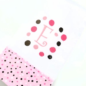 May include: White baby blanket with a pink polka dot border. The center features an embroidered pink letter "E" surrounded by pink and brown polka dots. A soft, cozy design.