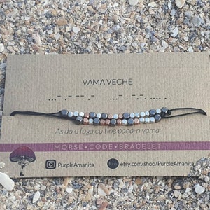 Peut inclure: Une carte marron avec le texte "VAMA VECHE" et un bracelet en cordon noir avec des perles argentées, noires et cuivrées. Le bracelet est un bracelet en code morse avec le message "As da o fuga cu tine pana-n vama". La carte comprend également le texte "MORSE CODE BRACELET" et le nom de profil sur les réseaux sociaux "PurpleAmanita".