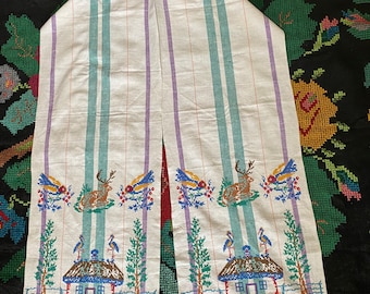 May include: A white linen tea towel with embroidered designs of birds, deer, houses, and flowers. The towel has green, blue, and purple stripes.