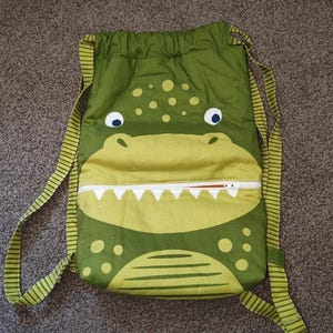 May include: A green drawstring backpack featuring a stylised crocodile face design. The bag includes a white horizontal zip across the centre that resembles teeth, with dark green and light green circular spots and striped shoulder straps.