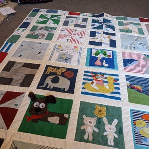 Può includere: Una trapunta patchwork con una varietà di quadrati colorati che presentano diversi temi di animali e libri per bambini. I quadrati sono cuciti insieme con tessuto bianco e la trapunta è bordata con un bordo bianco.