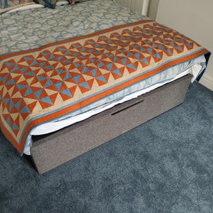 May include: A grey upholstered bed frame with a storage drawer. The bed is made with a white sheet and a blue and orange patterned quilt.