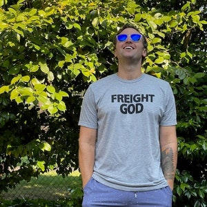 以下が含まれることがあります： グレーのTシャツに「FREIGHT GOD」という文字がプリントされたものを着ている人。青と白の模様のショートパンツと青いサングラスもかけています。