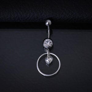 Könnte beinhalten: Silberner Bauchnabelring mit einem klaren Kristall und einem baumelnden Herz-Anhänger.