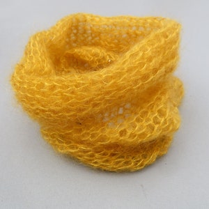 May include: A yellow knitted scarf made of a soft, fuzzy yarn. The scarf is loosely woven and has a delicate, airy texture.