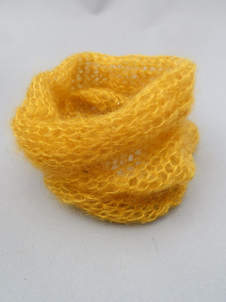 May include: A yellow knitted scarf made of a soft, fuzzy yarn. The scarf is loosely woven and has a delicate, airy texture.