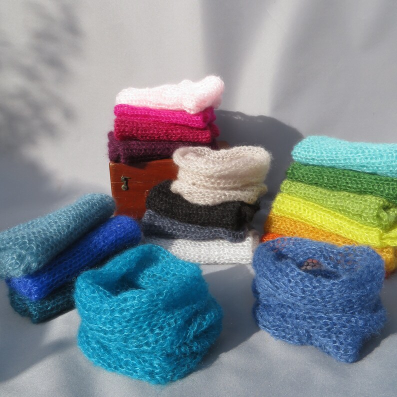 May include: A collection of colorful knitted scarves in various shades of blue, green, pink, and purple. The scarves are made of a soft, delicate yarn and are arranged in a pile on a white surface.