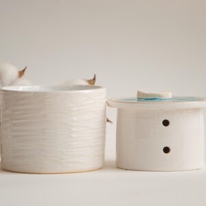 May include: Two white ceramic containers. One is a cylindrical jar with a textured surface. The other is a small, lidded container with two small holes and a blue and white design on the lid. Cotton bolls are in the background.