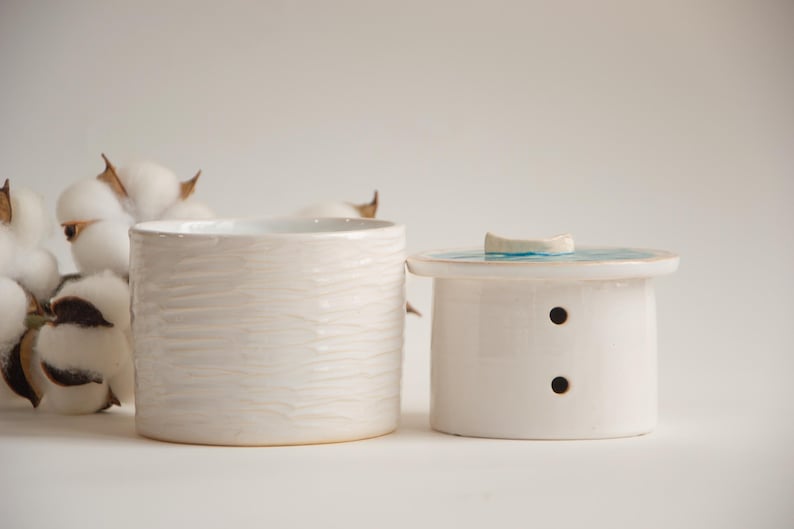 May include: Two white ceramic containers. One is a cylindrical jar with a textured surface. The other is a small, lidded container with two small holes and a blue and white design on the lid. Cotton bolls are in the background.