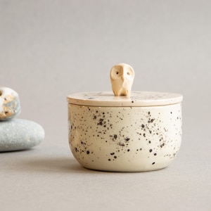 May include: A round ceramic jar with a cream-colored glaze and dark brown speckles. The lid features a small, sculpted owl figurine as a handle. The jar is accompanied by three smooth, rounded river stones placed to the left of the container.