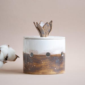 May include: A ceramic jar with a white top and a brown, speckled base. The jar has a lid and a decorative flower-shaped handle. Three circular holes are visible on the side. Fluffy cotton bolls are in the background.