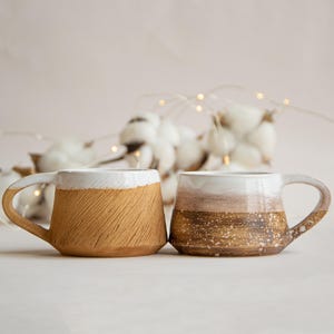 May include: Two ceramic mugs with a white glaze and brown and beige patterns. The mugs have a rounded shape and a handle on the side.