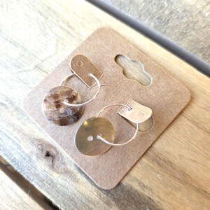 May include: A pair of hoop earrings with a button-like design. One earring is a light brown color, and the other is a gold color. The earrings are attached to a brown cardboard display card with a small hole for hanging.