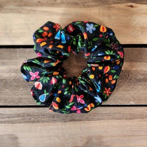 May include: A black scrunchie with a colorful floral pattern. The pattern includes red, orange, yellow, green, blue, and purple flowers and leaves.