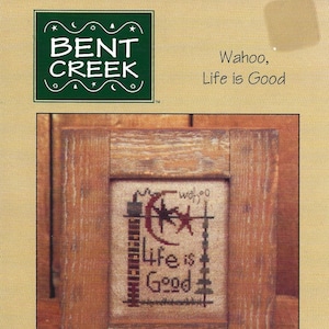 May include: A cross-stitch pattern featuring a rustic wooden frame with a beige background. The cross-stitch design includes a crescent moon, stars, and the text "Wahoo, Life is Good". The pattern is labeled "BC406" and is from Bent Creek.