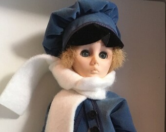 May include: A blue and black doll wearing a white scarf and holding ice skates. The doll has blonde hair and blue eyes. The doll is wearing a blue jacket with black buttons and black pants. The doll is holding a tag that says "Skater First Edition Currier & Ives Collection".