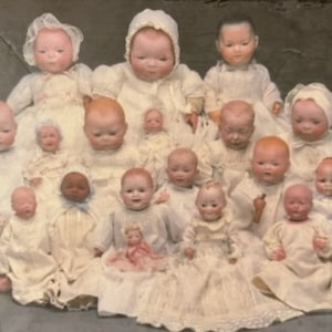 May include: A collection of antique porcelain dolls, some wearing bonnets and dresses, displayed in a group. The dolls are arranged in a variety of poses and sizes, with some sitting, standing, and lying down. The dolls are all dressed in white and have realistic features. The image is labeled "The Margaret Woodbury Strong Museum".