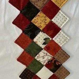 May include: A long table runner made of patchwork fabric squares in shades of red, green, brown, white, and yellow. The squares are sewn together in a diagonal pattern, creating a unique and eye-catching design.