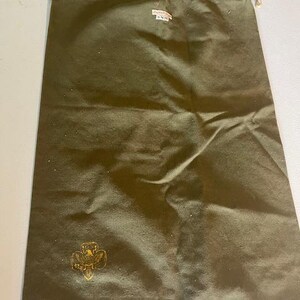 May include: An olive green drawstring bag with a gold emblem and a small tag. The bag is made of a durable fabric and has a drawstring closure with beige rope. The emblem appears to be a stylized floral design.