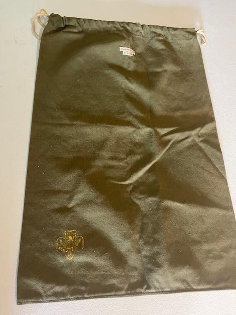 May include: An olive green drawstring bag with a gold emblem and a small tag. The bag is made of a durable fabric and has a drawstring closure with beige rope. The emblem appears to be a stylized floral design.