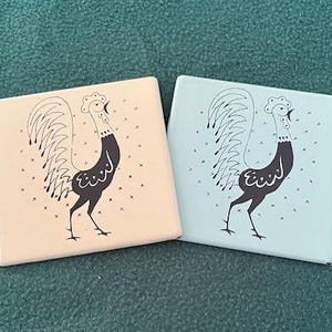 May include: Two ceramic rooster coasters, one in light pink and one in light blue. Both coasters feature a black rooster with a white tail and a white background with small black crosses.