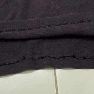 May include: Close-up of the bottom hem of a dark gray t-shirt. The hem is sewn with a zig-zag stitch and has a slightly frayed edge.