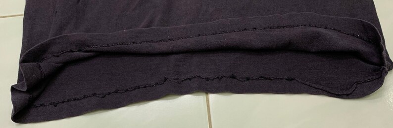 May include: Close-up of the bottom hem of a dark gray t-shirt. The hem is sewn with a zig-zag stitch and has a slightly frayed edge.