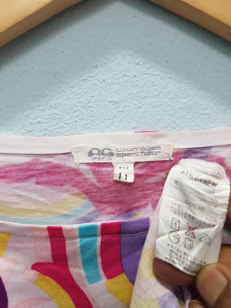 May include: A white, pink, yellow, blue, and purple patterned shirt with a white tag that says "Paris Sportwear" and "Size 11". The tag also has a black and white label with the text "ATN-0132 W" and "DOS 46".