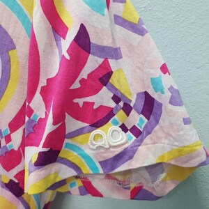 May include: A white t-shirt with a colorful abstract print. The print features pink, purple, yellow, and blue colors. The shirt has a white embroidered logo on the sleeve that says "QQ".