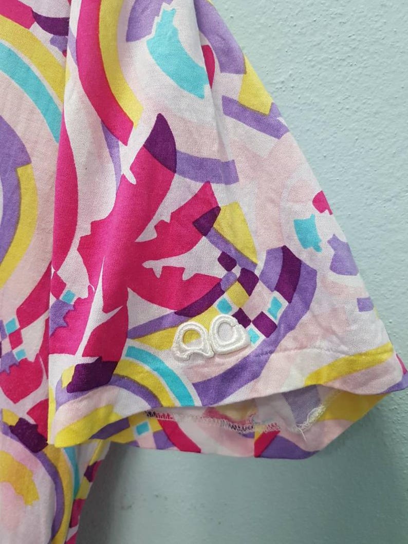 May include: A white t-shirt with a colorful abstract print. The print features pink, purple, yellow, and blue colors. The shirt has a white embroidered logo on the sleeve that says "QQ".