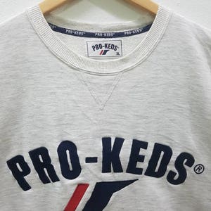May include: A white sweatshirt with a navy blue and red embroidered logo that says "PRO-KEDS" with a registered trademark symbol. The logo is arched and has a red and white stripe underneath it. The sweatshirt has a crew neck and is made of a soft, comfortable material.