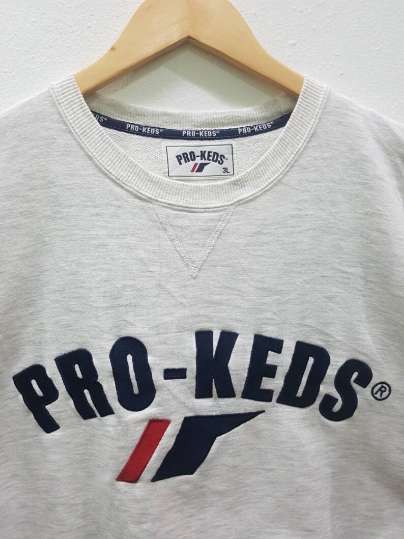 May include: A white sweatshirt with a navy blue and red embroidered logo that says "PRO-KEDS" with a registered trademark symbol. The logo is arched and has a red and white stripe underneath it. The sweatshirt has a crew neck and is made of a soft, comfortable material.