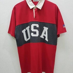 May include: Red, gray, and white striped short-sleeved polo shirt with the word "USA" in white letters on the gray stripe. The shirt has a white collar and a small American flag on the right sleeve.