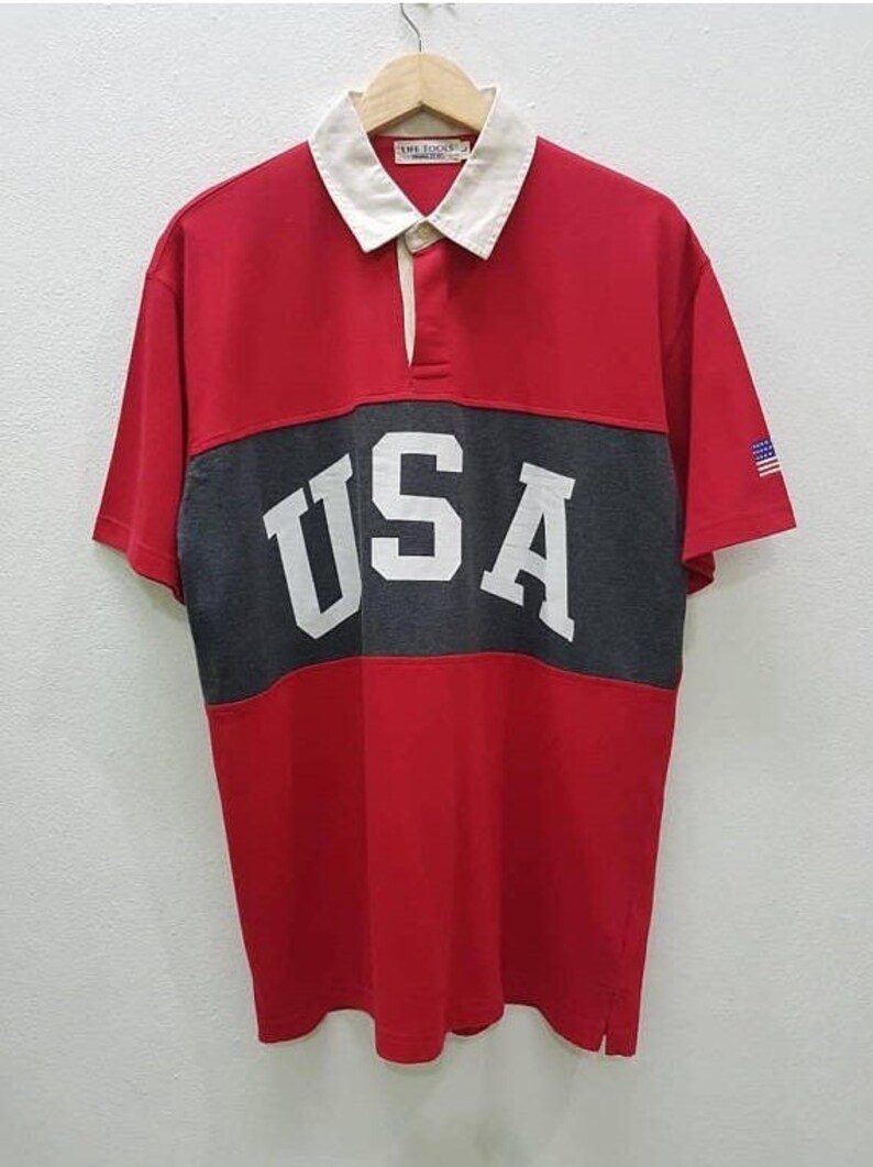 May include: Red, gray, and white striped short-sleeved polo shirt with the word "USA" in white letters on the gray stripe. The shirt has a white collar and a small American flag on the right sleeve.