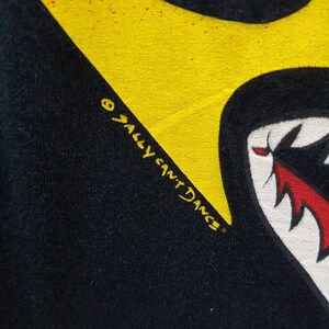 May include: Black t-shirt with a yellow and red shark tooth graphic. The text "Sally  Sant'Dng" is printed in yellow on the shirt.